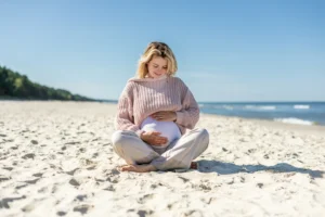 Kwas foliowy - właściwości, działanie, źródła, suplementy. Kobieta w ciąży siedzi na plaży w swetrze i długich spodniach, w tle widać morze.