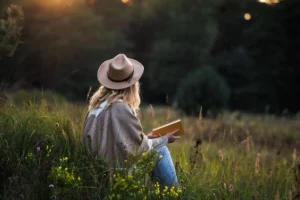 Kobieta w blond włosach, kapeluszu, jeansach i ponczo siedzi na trawie, na leśnej polanie z książką i praktykuje trening uważności mindfulness.